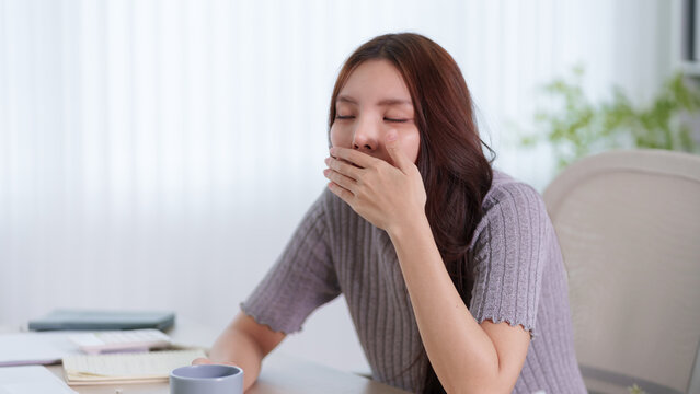 Young Asian woman yawning and covering mouth while sitting at home desk with notebook and coffee cup