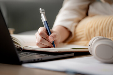 Fototapeta premium Online learning and homework study concept. Student writing notes while using a laptop at home, representing distance education, e-learning, self-study, online classes and academic success.