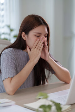 Young Asian woman covering face in distress while working on laptop at home desk with copy space
