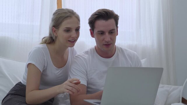 Caucasian couple sitting together on bed at home looking at laptop computer screen to browse online store website for shopping purchase order and internet video