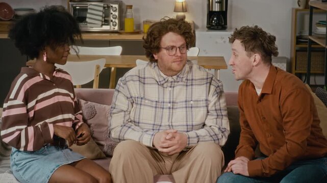 Multiethnic young friends sitting on sofa, talking and laughing together in cozy apartment. Clip is ideal for diversity marketing and youth lifestyle advertisements