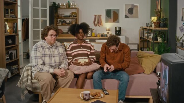 African American woman and Caucasian men sitting on sofa, eating popcorn, and watching television. Clip is suitable for home entertainment, streaming services, or lifestyle brand marketing