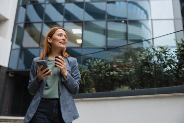 Fototapeta premium Businesswoman using her smartphone and drinking coffee outside an office building during a work break.