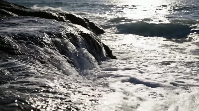 Ocean waves crashing against dark coastal rocks at sunset, serene seascape background, with white sea, foam and sparkling, sunlight reflections