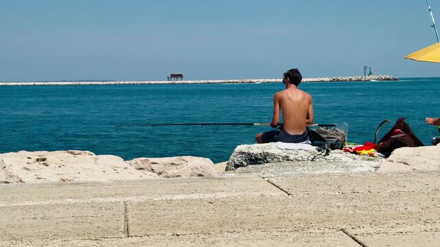 Gebr&auml;unter dunkler Junge angelt mit Schirm in der sonnigen Hafeneinfahrt von Chioggia
