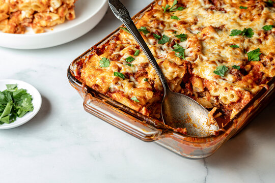 A baking dish of freshly made baked ziti hot out of the oven, with one serving.