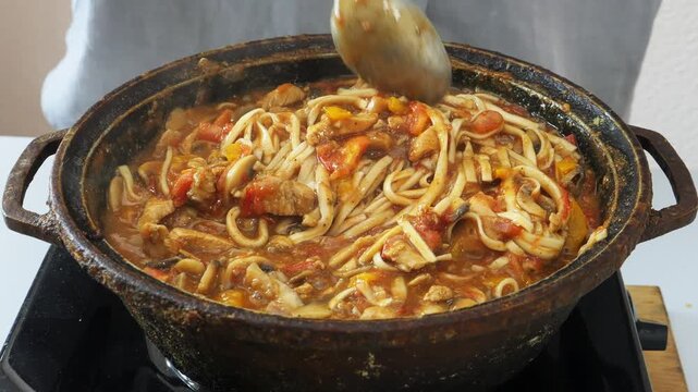 Vegetables with chicken and udon noodles are stewed in a frying pan in Pad Thai sauce and spices stirred with a spoon. Medium plan.