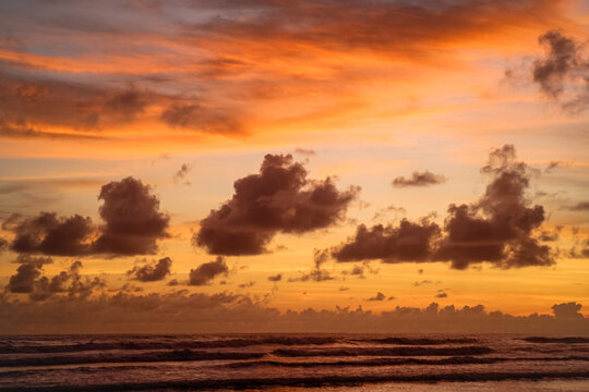 Beautiful seascape of rolling ocean waves under a vibrant orange and golden dramatic evening sky with scattered clouds at Parangtritis Beach, Indonesia.