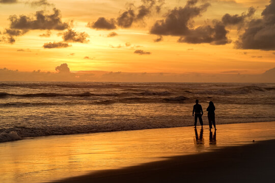 Silhouette of a couple walking along the shoreline of Parangtritis Beach, Yogyakarta during a beautiful golden sunset with stunning water reflections.