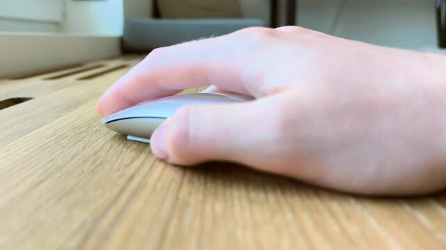 Hand on a mouse viewed from the thumb side. Fingers clicking on a wooden desk during computer use. Close-up angle of everyday digital navigation..