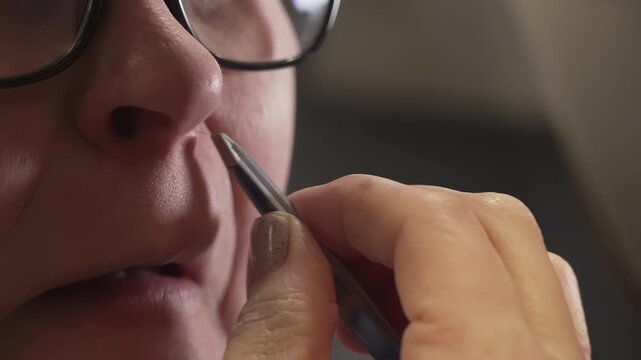 Close-up of a woman using a grooming tool to remove nose hair, focusing on her hand and facial features, with a blurred background that emphasizes the personal care setting