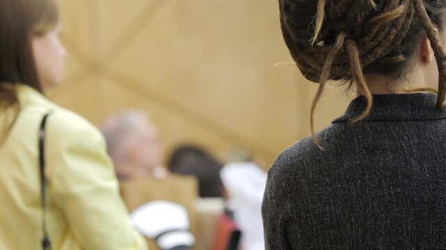 Two women stand in a conference hall. One has braided hair, wearing a dark blazer. The other wears a yellow jacket. Background shows seated attendees in warm lighting. Focus is on back views