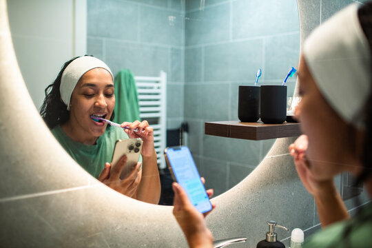 Woman brushing teeth while using smartphone in bathroom mirror