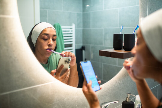 Woman brushing teeth and using smartphone in home bathroom mirror