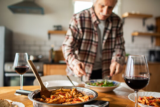 Senior man serving salad in cozy home kitchen with pasta and red wine