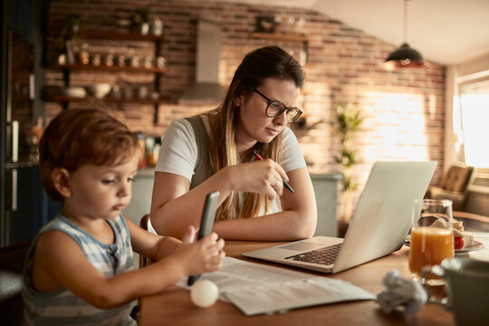 Mother working remotely on laptop at kitchen table with toddler