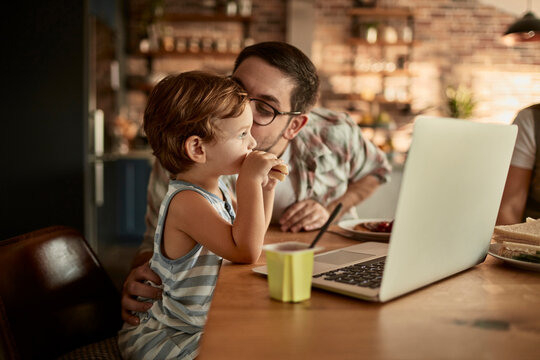 Father and toddler eating snack by laptop in home kitchen