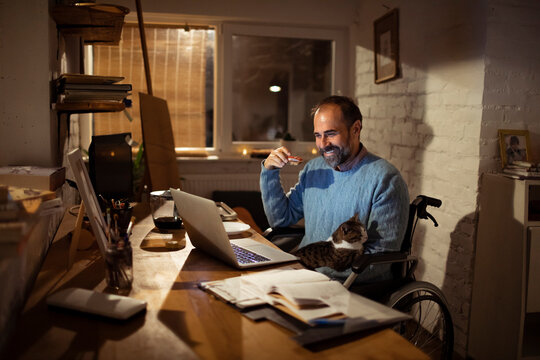 Man in wheelchair working from home office with cat at night