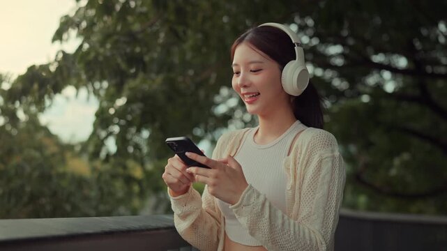 Happy young Asian woman wearing headphones using smartphone to listen to music online podcast in park, cheerful girl smiling while holding mobile phone outdoors leisure relaxed technology lifestyle.