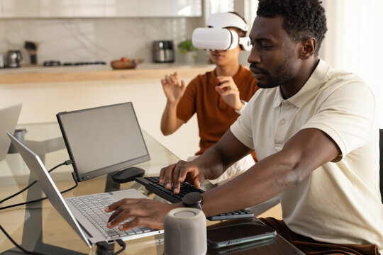 Diverse male coworkers working at glass dining table in kitchen, using laptop and VR headset