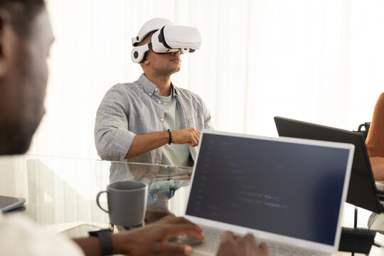 Diverse coworkers watching man wearing white VR headset gesturing over laptops at conference table