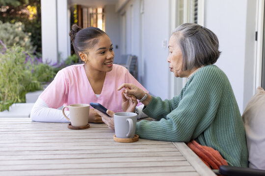 Senior Asian woman and African American nurse sitting on porch, pointing smartphone, holding mugs