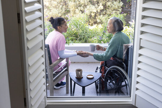 Senior Asian woman and female caregiver holding hands, sipping mugs on porch with wheelchair