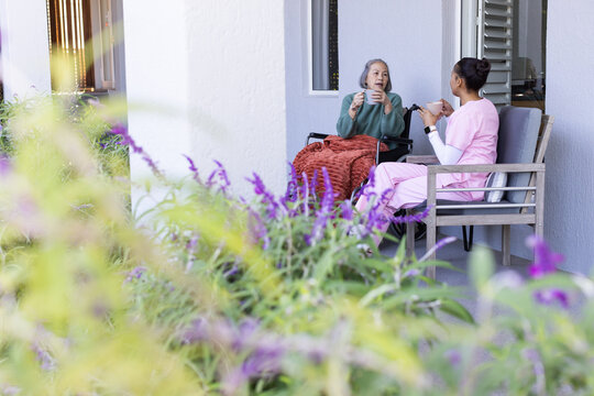 Senior Asian woman sitting in wheelchair holding mug, caregiver in pink scrubs on porch, copy space