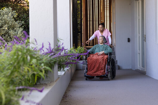 Asian senior sitting in wheelchair with rust throw, pink-scrub caregiver pointing at purple flowers