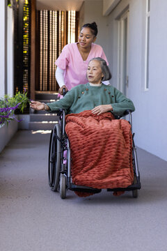 Senior woman and caregiver walking down corridor pushing wheelchair with rust blanket, pink scrubs