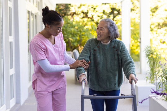 African American caregiver scrubs holding Senior Asian woman in green sweater using walker on porch