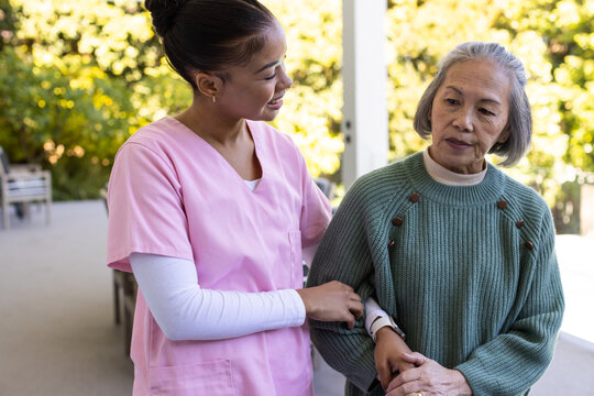 Senior Asian woman and African American female nurse helping on patio in green sweater, pink scrubs