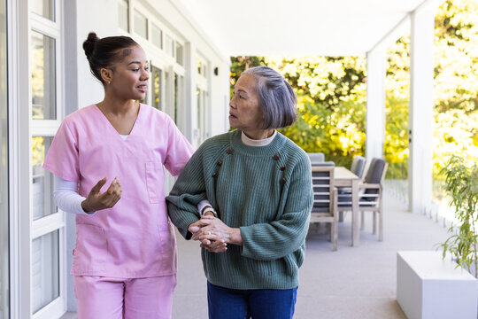 Diverse female caregiver and Asian senior walking on porch helping green sweater watch pink scrubs