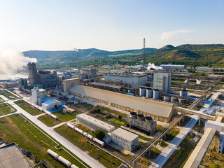 Fototapeta premium Smoke rises from a large industrial plant surrounded by green hills during the early afternoon hours
