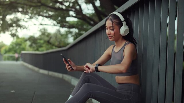 Fit Asian woman sitting on city park bridge looking at smartwatch and holding smartphone after running exercise, sporty girl wearing headphones checking fitness workout data and heart rate