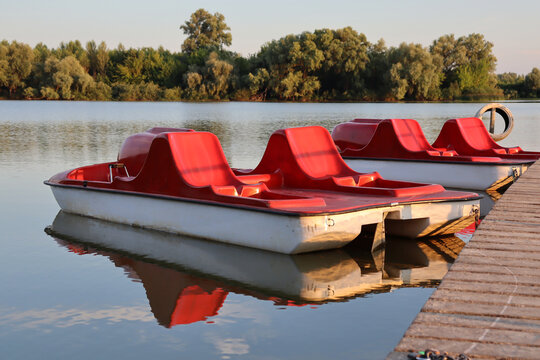 Red pedal boats sit by the water in the late afternoon sun near a tree-lined shore