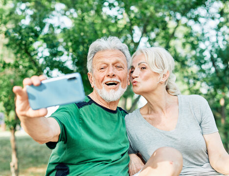 Happy active senior couple having fun using smartphone, taking selfie, or having a video call and wearing sportswear, after having an exercise sport activity outdoors