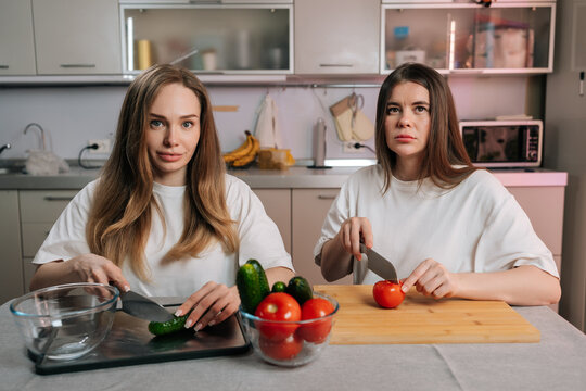 Two young women sit at kitchen table, preparing fresh salad. One smiles while cutting cucumbers, other looks unhappy cutting tomatoes. Concept of different attitudes towards cooking or dieting.
