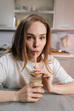 Vertical portrait of young caucasian woman enjoying healthy smoothie at home. She sips from glass with eco friendly straw, looking at camera. Soft light, shallow depth of field, wellness concept.