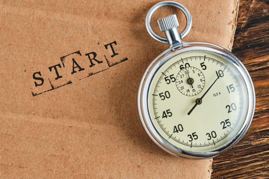 Horizontal view metal stopwatch lying next to stamped START word on cardboard and dark wood.