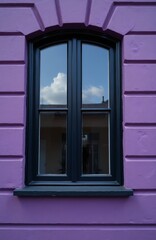 Obraz premium Black window frame installed in vivid purple wall. Reflection of bright blue sky with white clouds visible in glass. Vintage house exterior shows architectural design. Vibrant paint color on old
