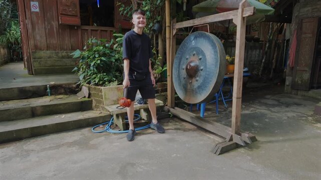 Young man striking traditional Buddhist temple gong after making silent wish, Male traveler taking part in sacred spiritual ritual at peaceful Asian monastery courtyard 4k