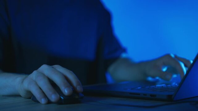 close-up shot of a person's hand using a computer mouse and laptop keyboard in a dark room illuminated by cool blue lighting. 