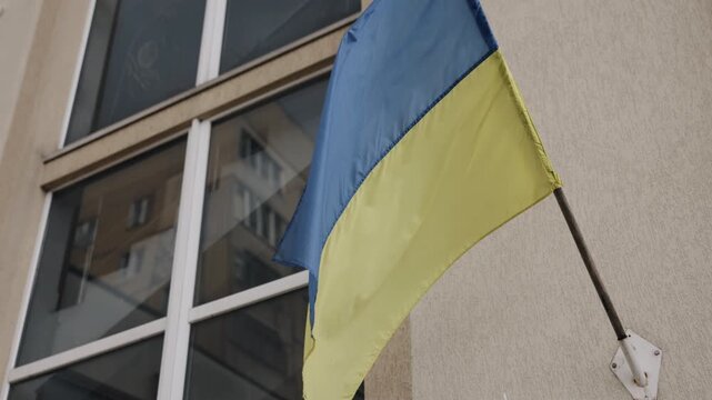 Ukraine flag, travel destination, support Ukraine. Blue and yellow Ukrainian national flag hangs on beige institutional building, with glass windows forming background.