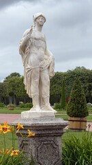 Obraz premium Classical white marble statue of a male figure standing on a granite pedestal in a manicured summer park garden.