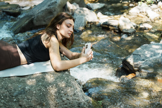 Young woman capturing beautiful nature with her phone by a mountain stream. Mobile photography, outdoor adventure, authentic travel moments and digital connection.