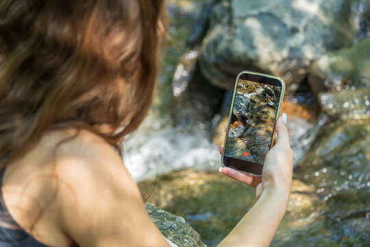 Young woman recording nature video on her phone by mountain stream. Authentic content creation, travel storytelling, sharing outdoor experiences and enjoying beauty of nature