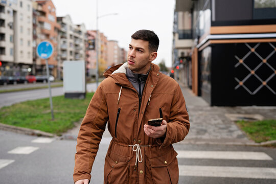 A stylish young man wearing a brown parka jacket holds a mobile phone while crossing an urban road, checking navigation apps or waiting for a ride share on an overcast autumn afternoon.