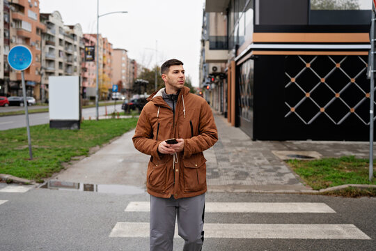 A stylish pedestrian stands near a crosswalk in an urban neighborhood, looking aside while holding a mobile phone during a cool autumn morning.