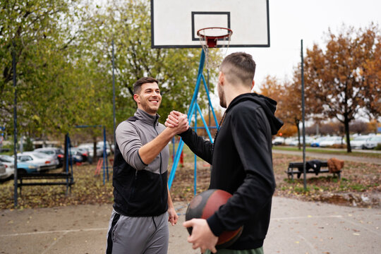 Athletic young male friends greet each other with a strong handshake after a match on a streetball court during autumn.